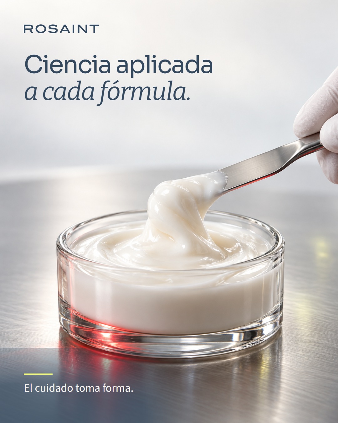Escena conceptual de formulación: una mano enguantada levanta una textura blanca con una espátula sobre un recipiente de vidrio.